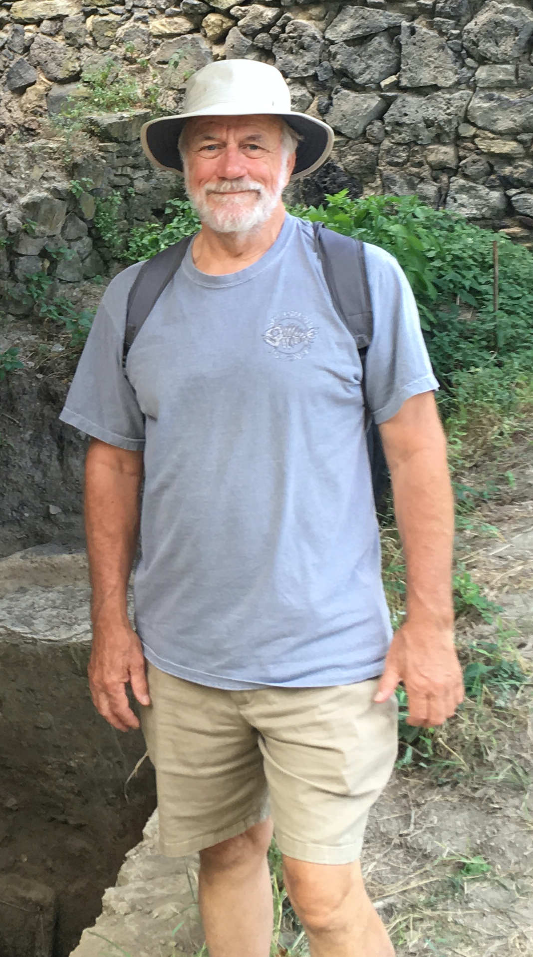Dr. Peter Olver standing outside wearing white hat, blue t-shirt, khaki shorts, and backpack. 
